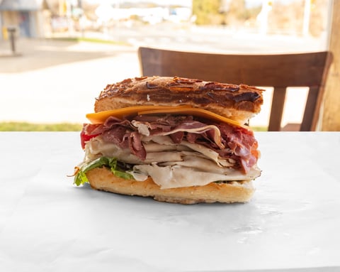 Gourmet sandwich stacked with prosciutto, turkey, cheese, and greens on a crusty roll, served on a white plate outdoors