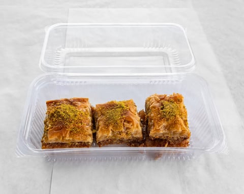 Three square baklava pastries with green pistachio topping in a clear plastic takeout container on white surface