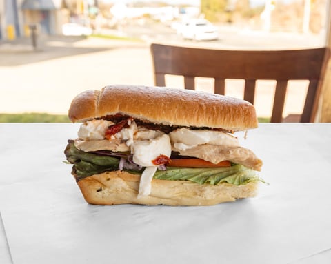 Gourmet sandwich on a toasted bun with chicken, melted cheese, fresh greens, and roasted vegetables on a white plate